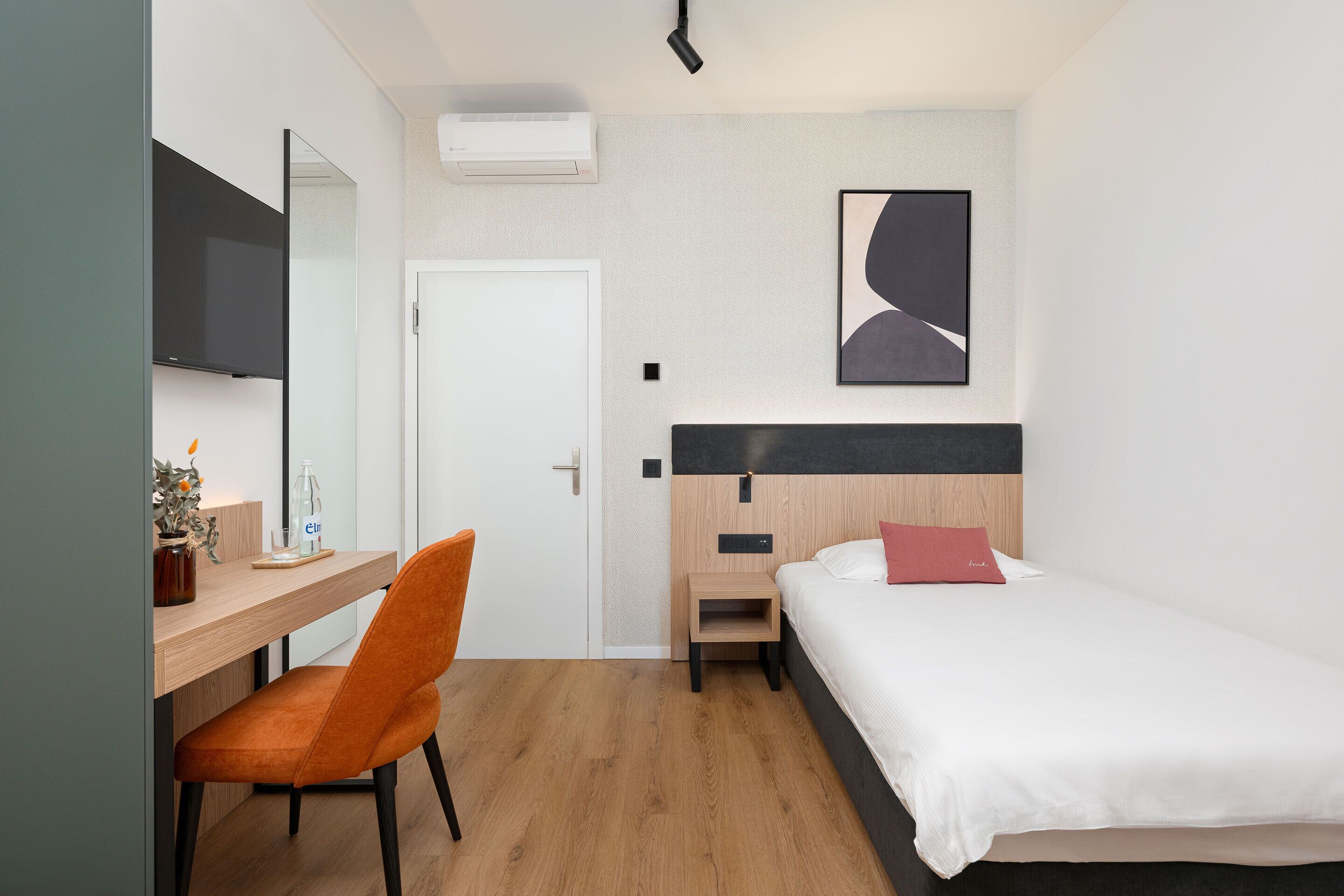 Ein modernes Hotelzimmer mit einem Holztisch, einem Stuhl, einem Bett mit weißer Matratze und einem roten Kissen, einer Wand-Klimaanlage und einem gerahmten abstrakten Kunstwerk.