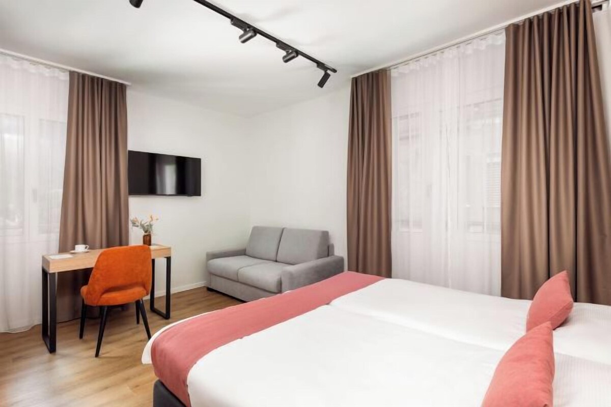 A hotel room with a bed, a desk, a chair, a sofa, and a TV.