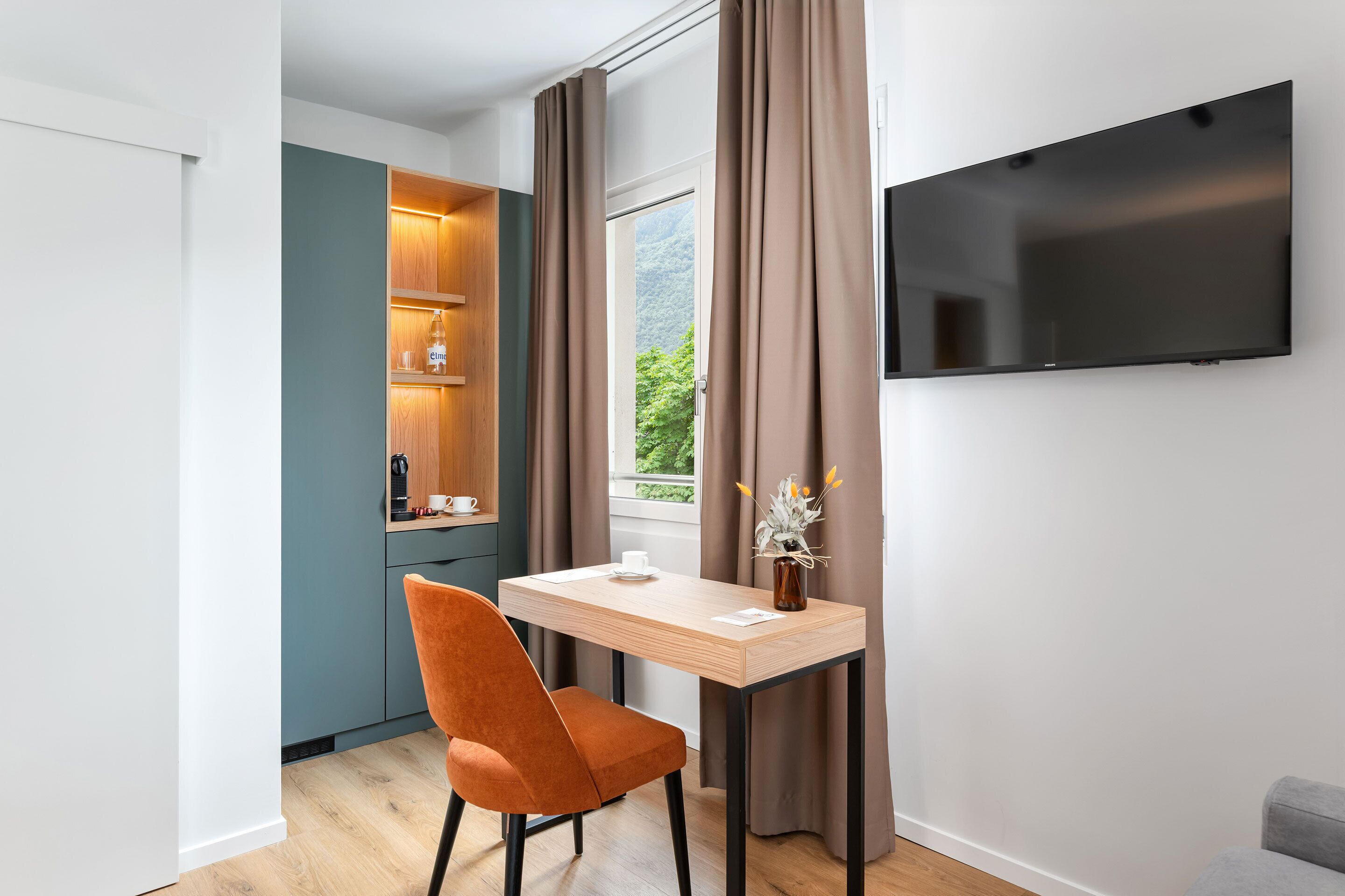 Quarto moderno com escrivaninha de madeira, uma cadeira, televisão e janela com cortinas.