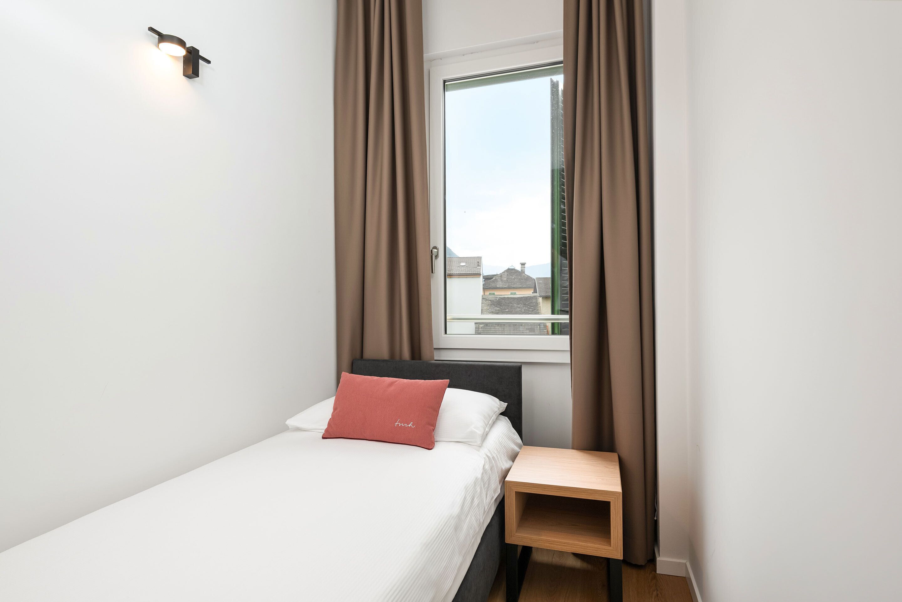 Une petite chambre à coucher, bien rangée, avec un lit simple, une table de chevet et une fenêtre avec des rideaux.