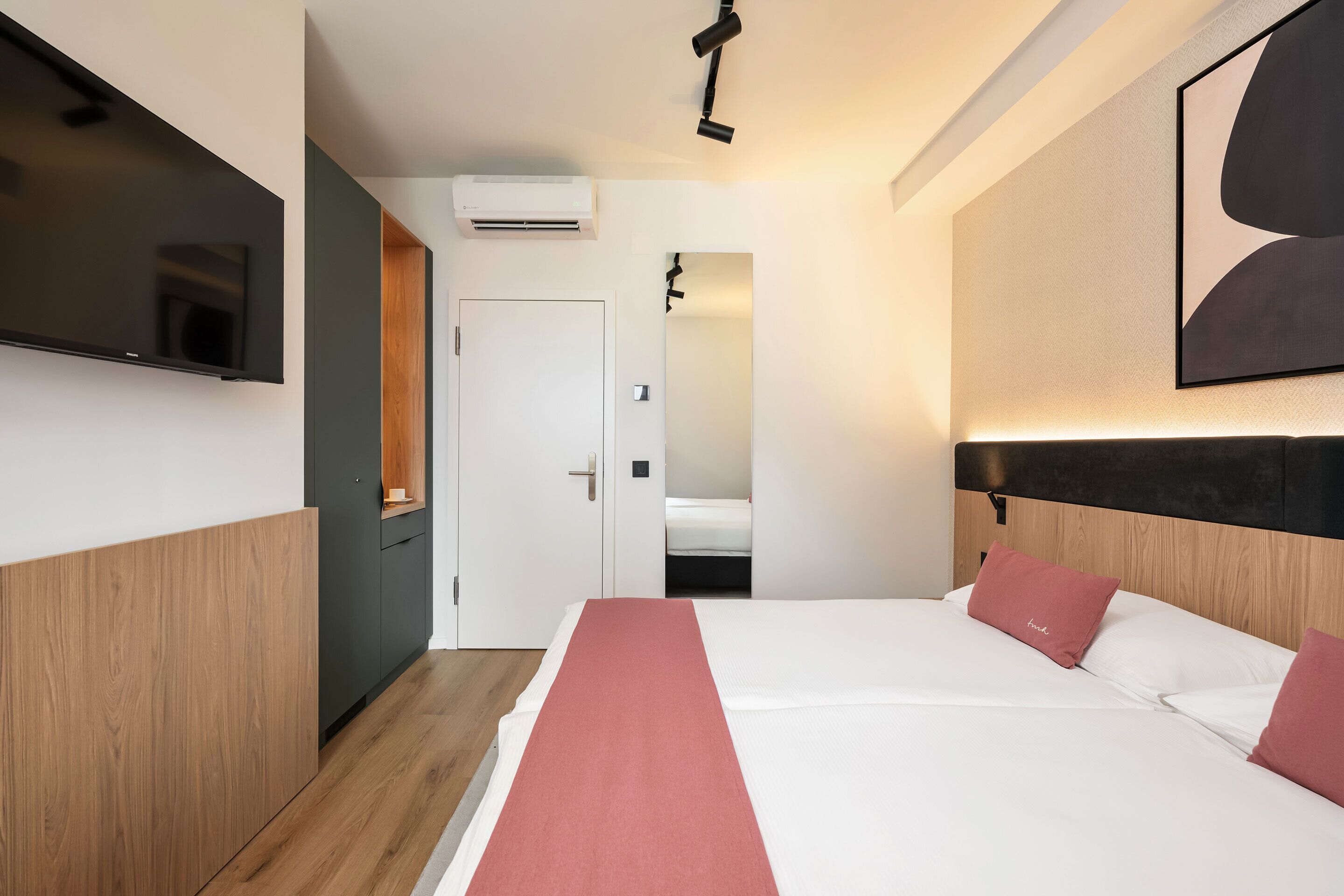 A modern hotel room with a bed, a flat-screen TV, an air conditioning unit, and a door leading to another room.