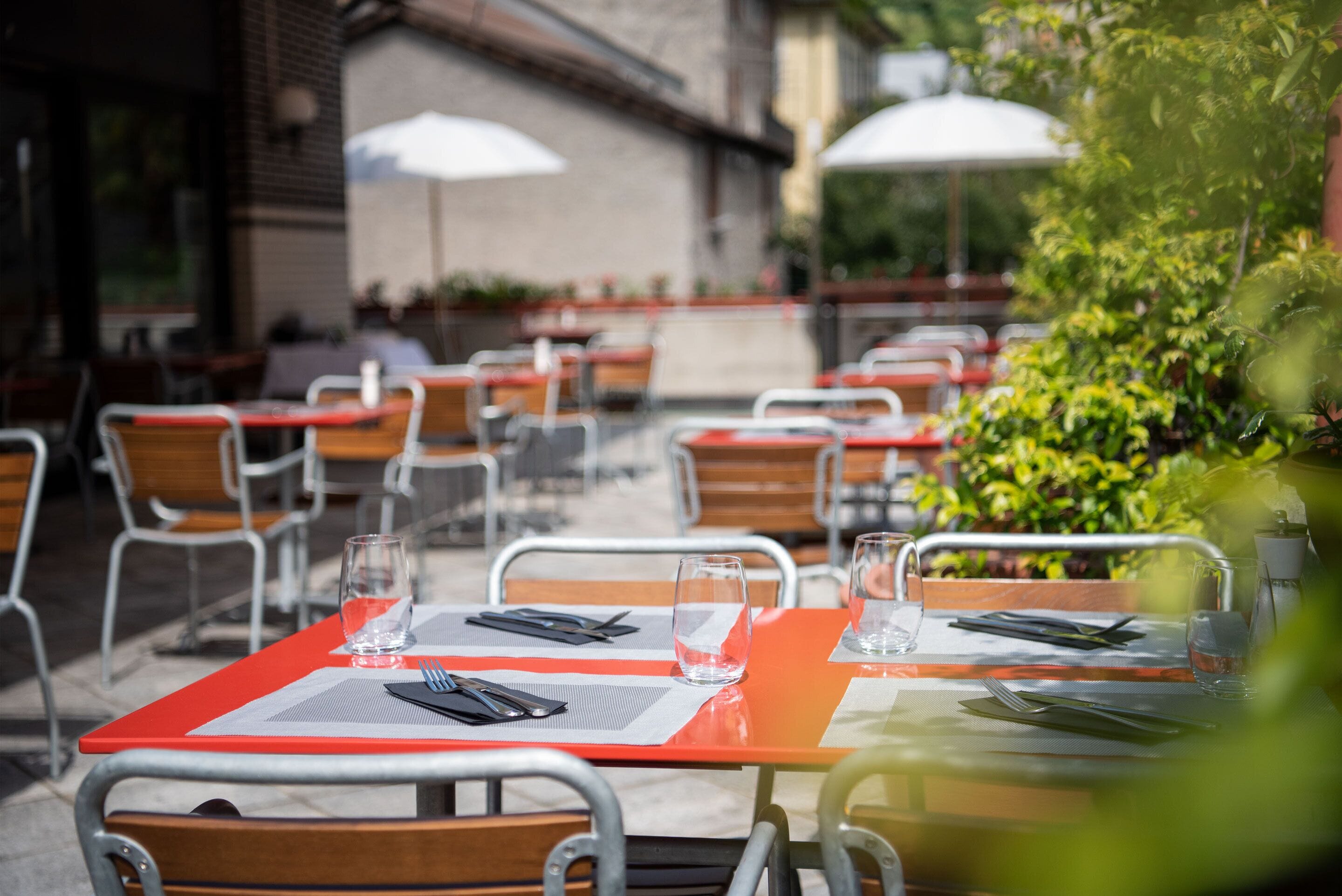 Des tables à l’extérieur, accompagnées de chaises en métal, dont certaines sont dotées de parasols, et un bâtiment en briques en arrière-plan.
