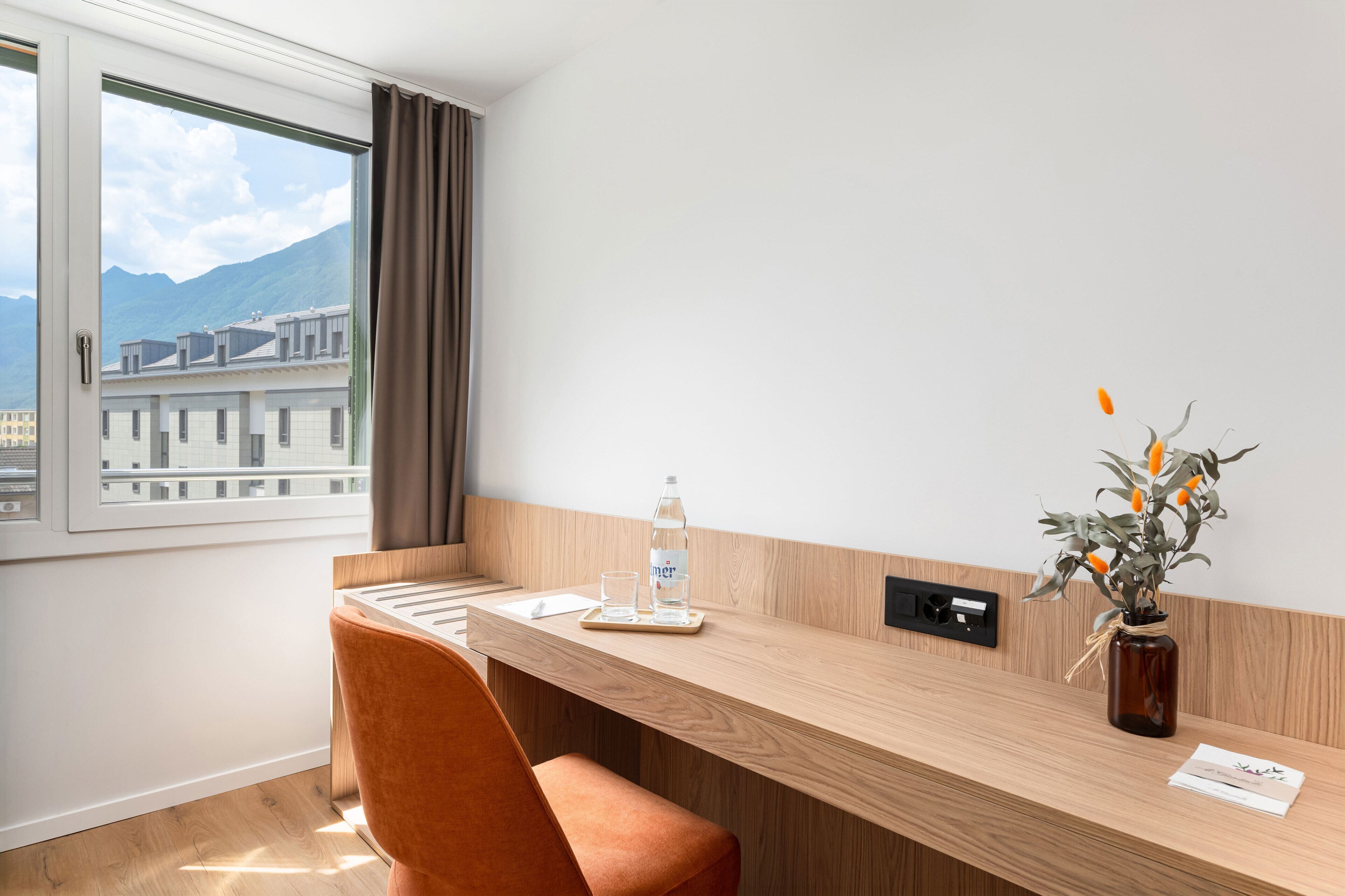 Une chambre d’hôtel avec un bureau en bois, une chaise, une bouteille d’eau, un verre, un vase avec des fleurs et une vue sur les bâtiments et les montagnes par la fenêtre.