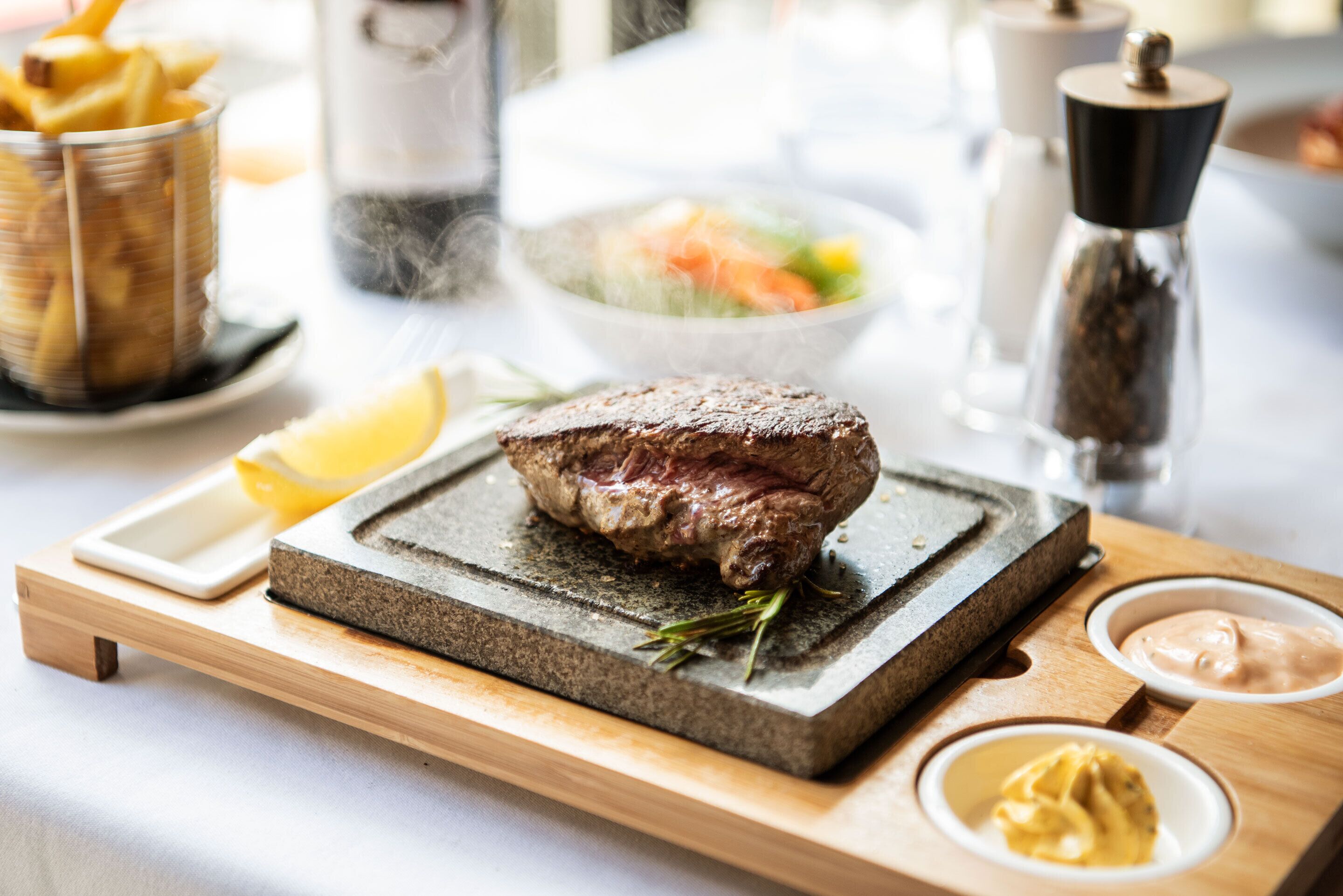 A steak on a stone board with condiments and a salad in the background.