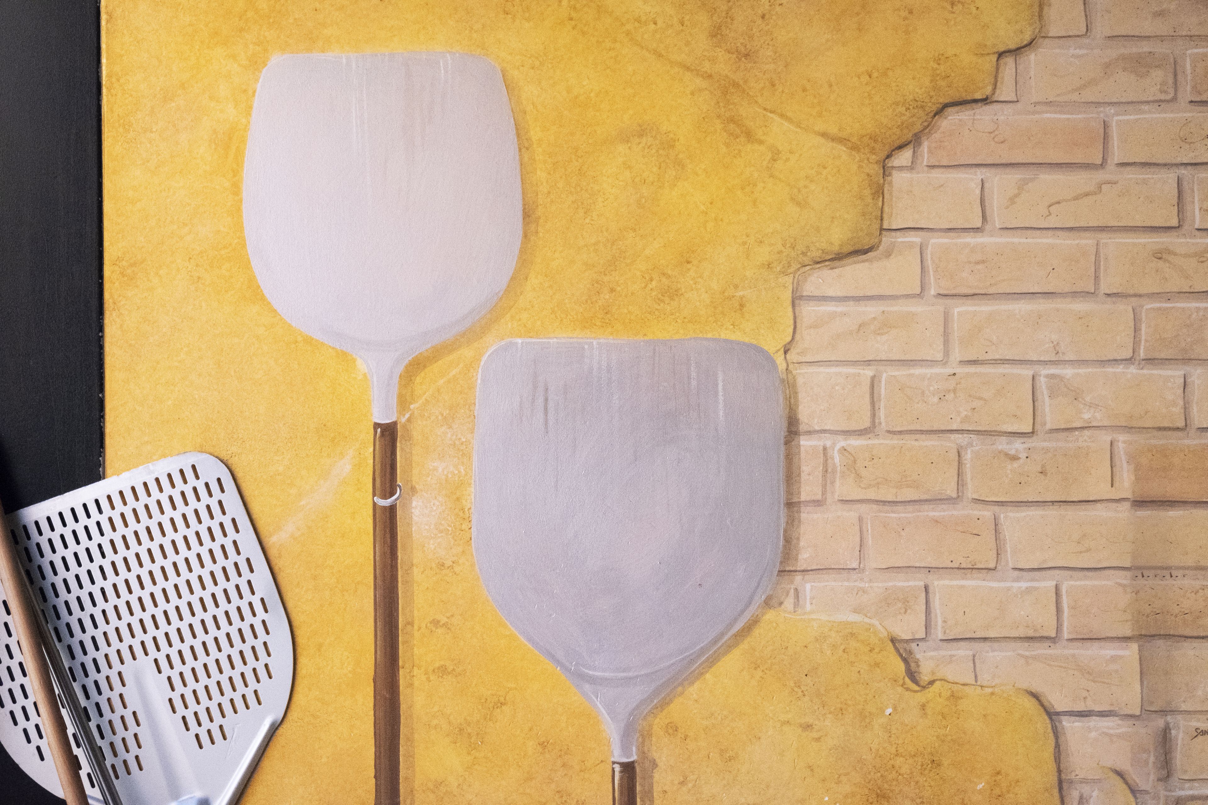 Deux ustensiles de cuisine sont posés contre un mur jaune orné d’un motif en briques.