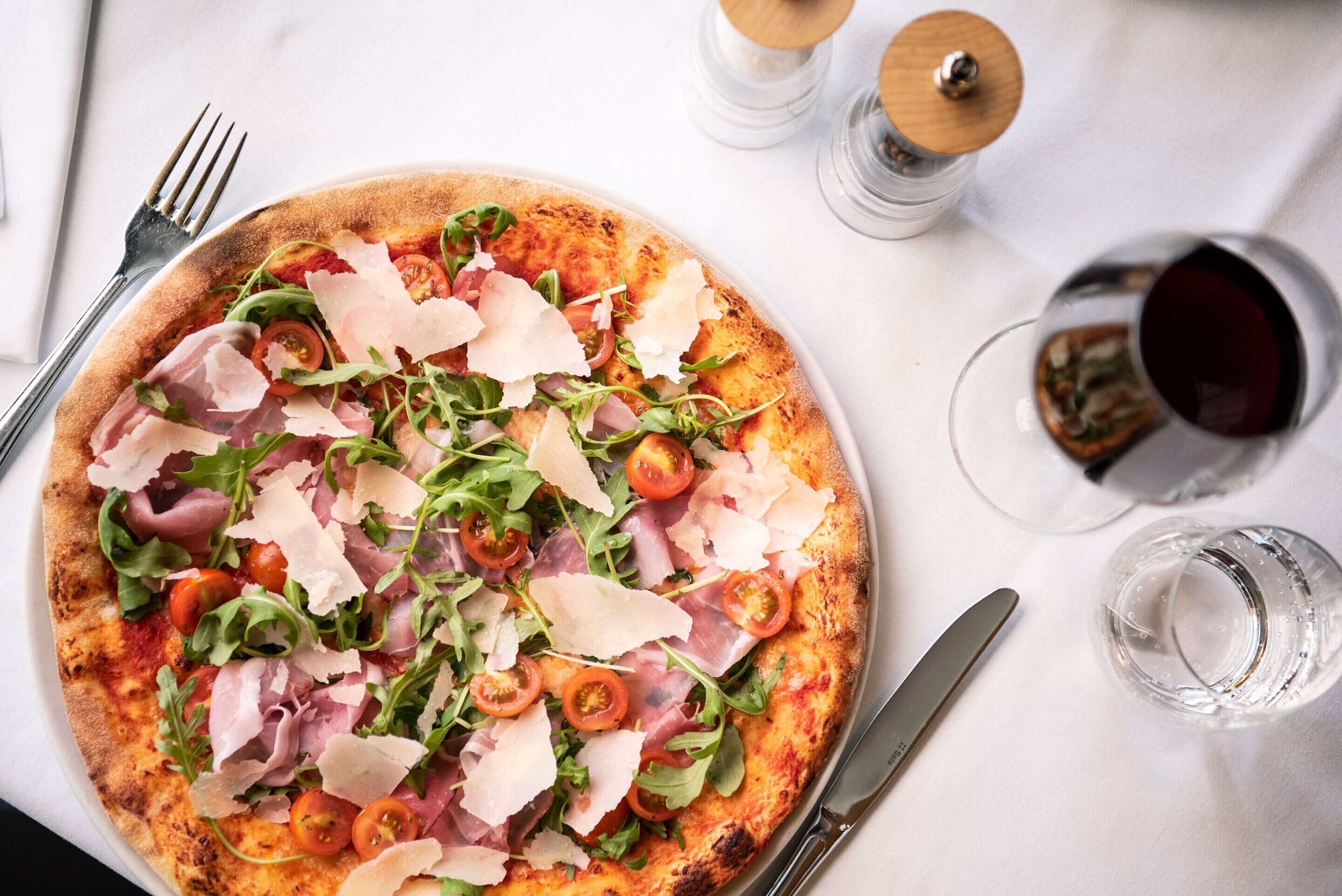 A pizza with various toppings on a white plate, accompanied by a fork, knife, and two glasses of red wine on a white tablecloth.