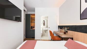 Habitación de hotel con cama, escritorio, silla, televisor y baño visible a través de una puerta abierta.