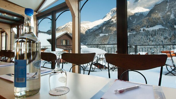 A table with a bottle, a glass, and a pen, overlooking a snowy mountain landscape. - Hotel Suisse (Champery)