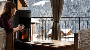 A person sitting at a wooden table with a glass of drink, a book, and a remote control. The table is near a black wood-burning stove. - Hotel Suisse (Champery)