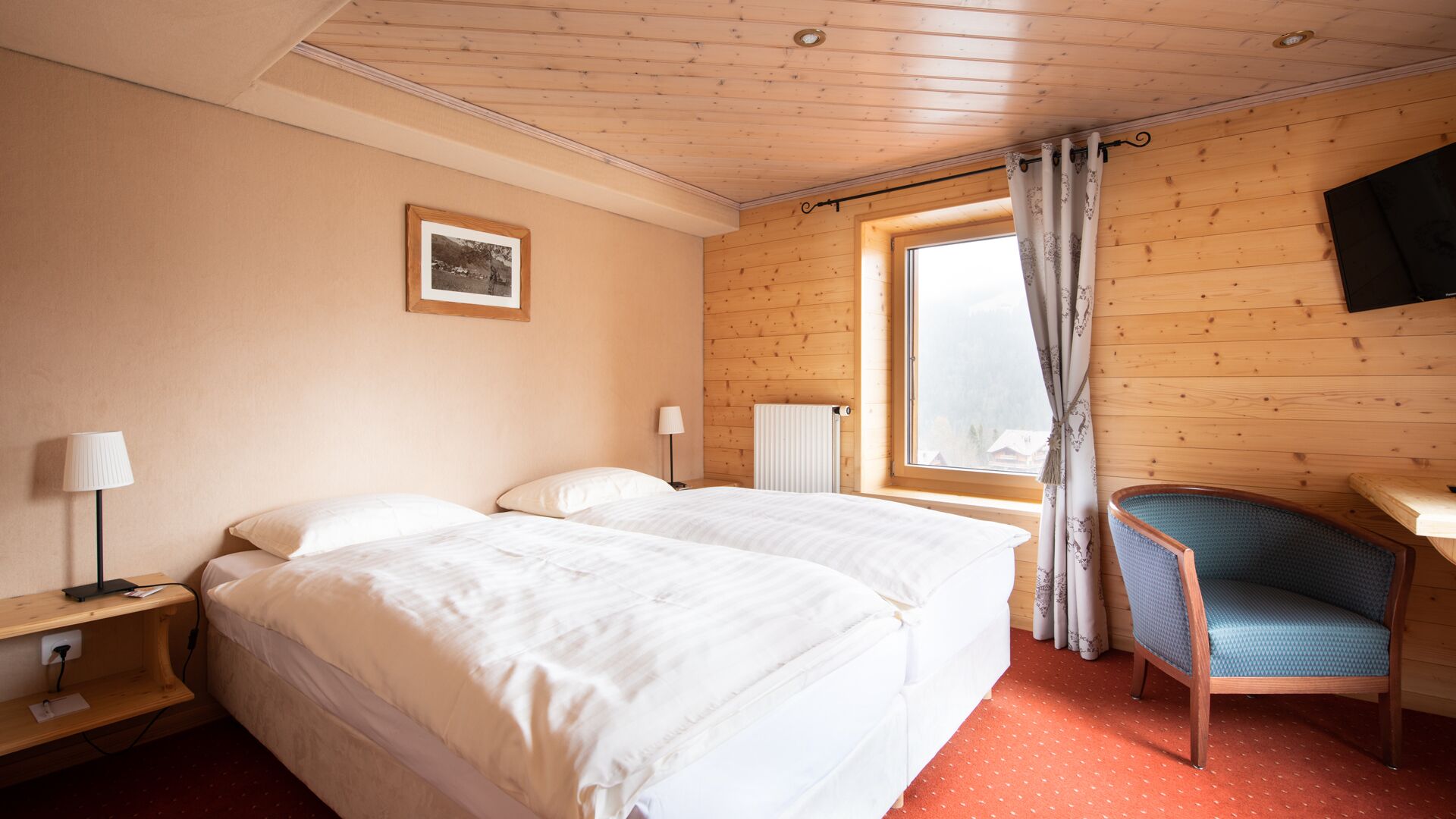 Quarto com duas camas, janela com cortinas, uma cadeira e televisão fixada na parede.