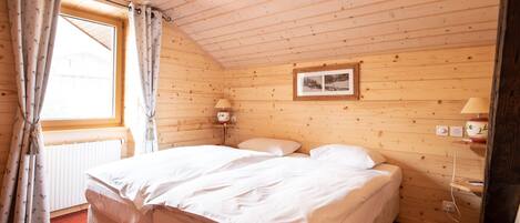 Una habitación de madera con una cama, un ventanal con cortinas, un cuadro en la pared y una mesita de noche con una lámpara.
