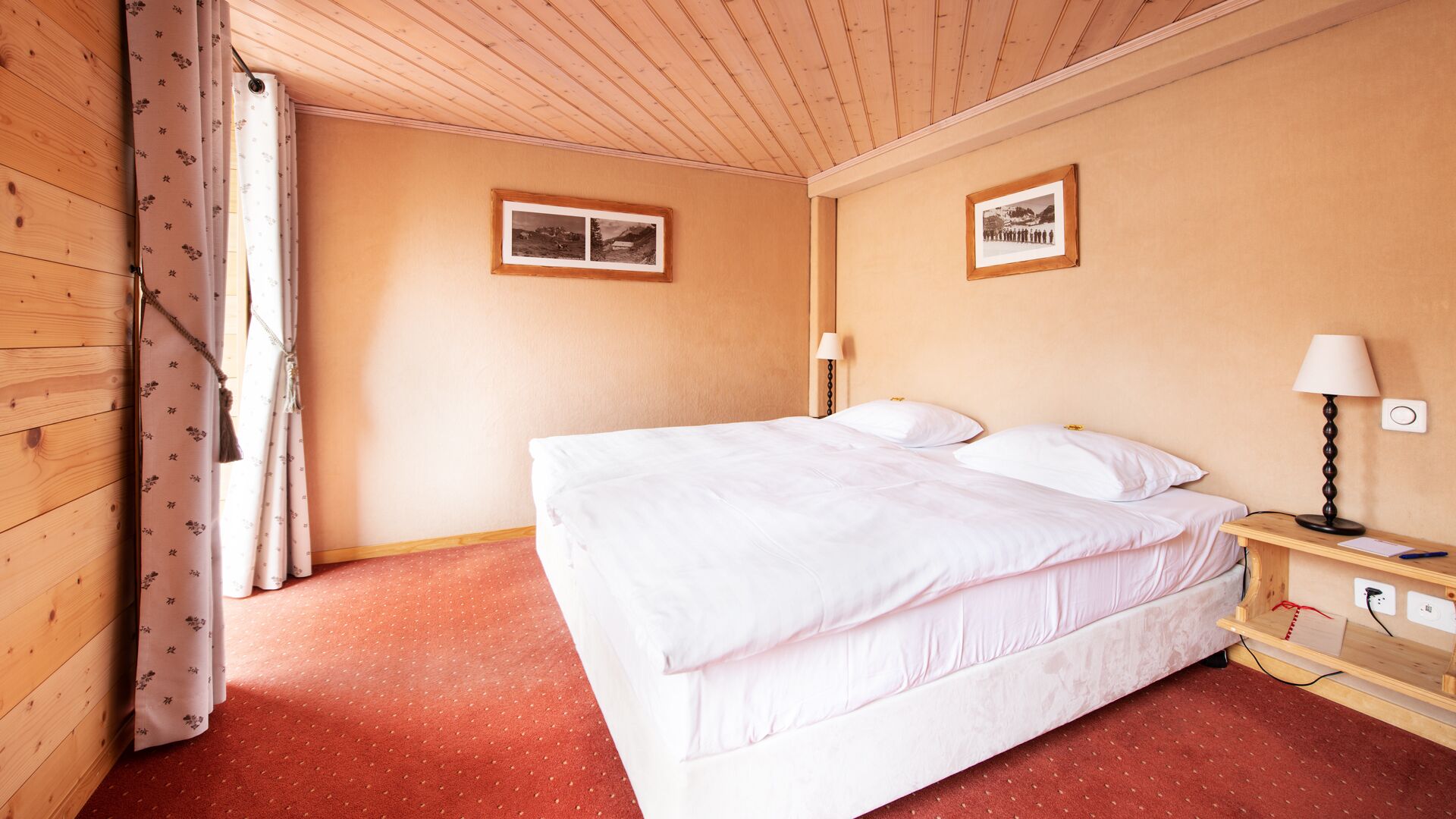 A hotel room with a wooden ceiling, a bed with white bedding, a nightstand with a lamp, and two framed pictures on the walls.