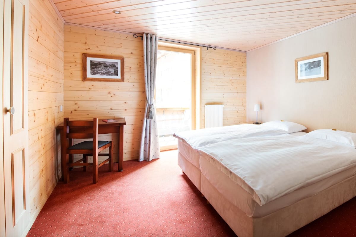 A room with a wooden floor, a bed with white bedding, a desk with a chair, and two framed pictures on the walls.