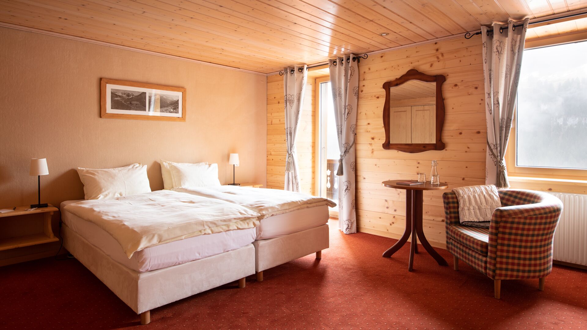 A cozy hotel room with a double bed, a small table with a chair, a mirror, and a window with curtains.