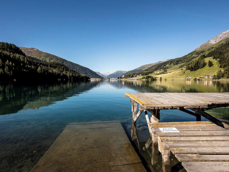 A wooden dock extending into a calm lake surrounded by forested mountains.