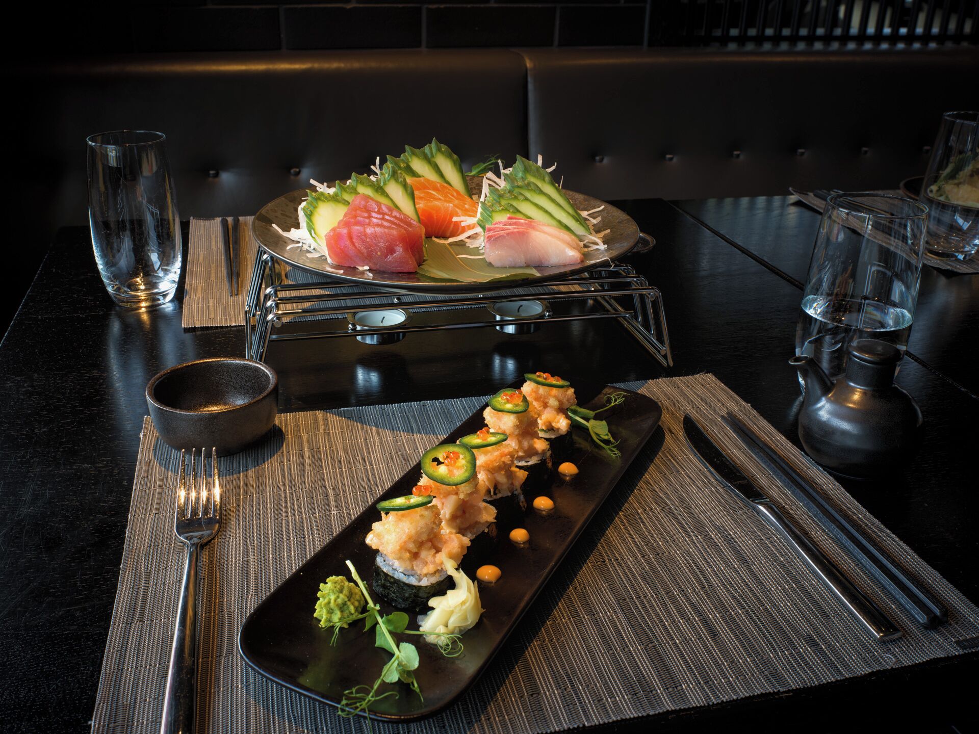 Una bandeja negra con un plato de sushi que incluye varios tipos de sushi y sashimi, acompañado de un recipiente pequeño con salsa y un vaso de agua.