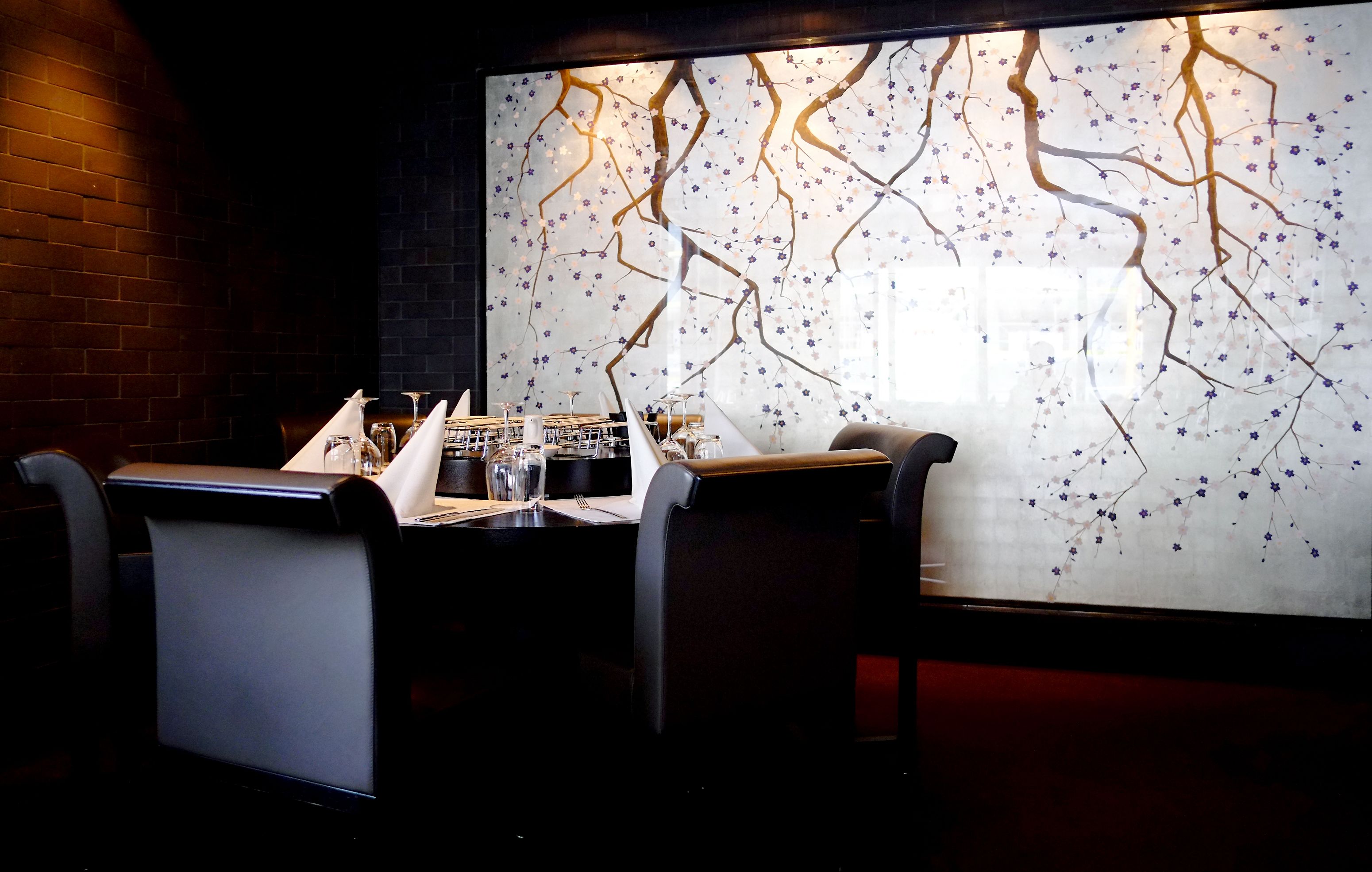 Interior de un restaurante con pared de ladrillo, una larga mesa dispuesta con servilletas blancas, vasos y una carta.