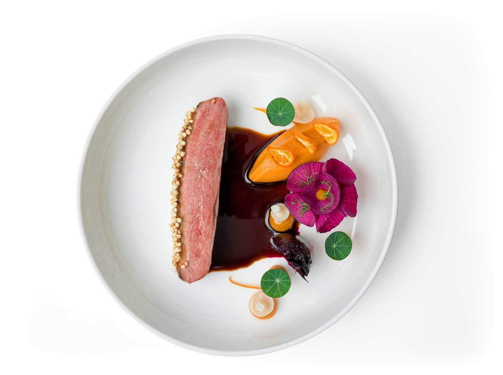 A white plate with a dish consisting of a slice of meat, a slice of orange, a purple flower, and some green leaves.