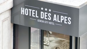 L’extérieur de l’hôtel Des Alpes Geneva City se distingue par son design moderne, ses grandes fenêtres et un pot de fleurs placé devant l’entrée.