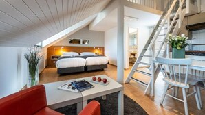 Une chambre d’hôtel moderne avec un espace nuit en mezzanine, un coin repas avec une table et des chaises blanches, et une cuisine visible au fond.