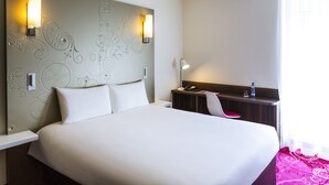 Habitació d'hotel amb un llit gran, dues làmpades penjades a la paret, un escriptori amb una cadira i una cambrera de nit amb un telèfon i una ampolla d'aigua.