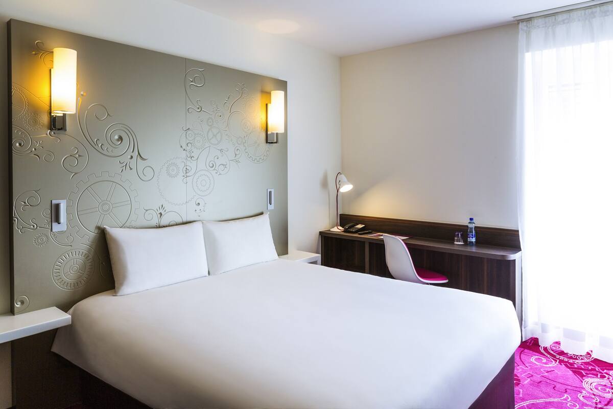 Ein Hotelzimmer mit einem großen Bett, zwei Wandlampen, einem Schreibtisch mit Stuhl sowie einem Nachttisch mit Telefon und Wasserflasche.