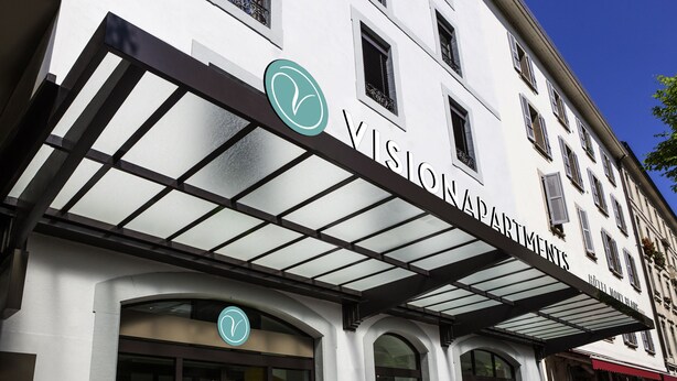 Un hotel anomenat “VISION APARTMENTS” amb una façana moderna, grans finestres i un logotip ben visible.