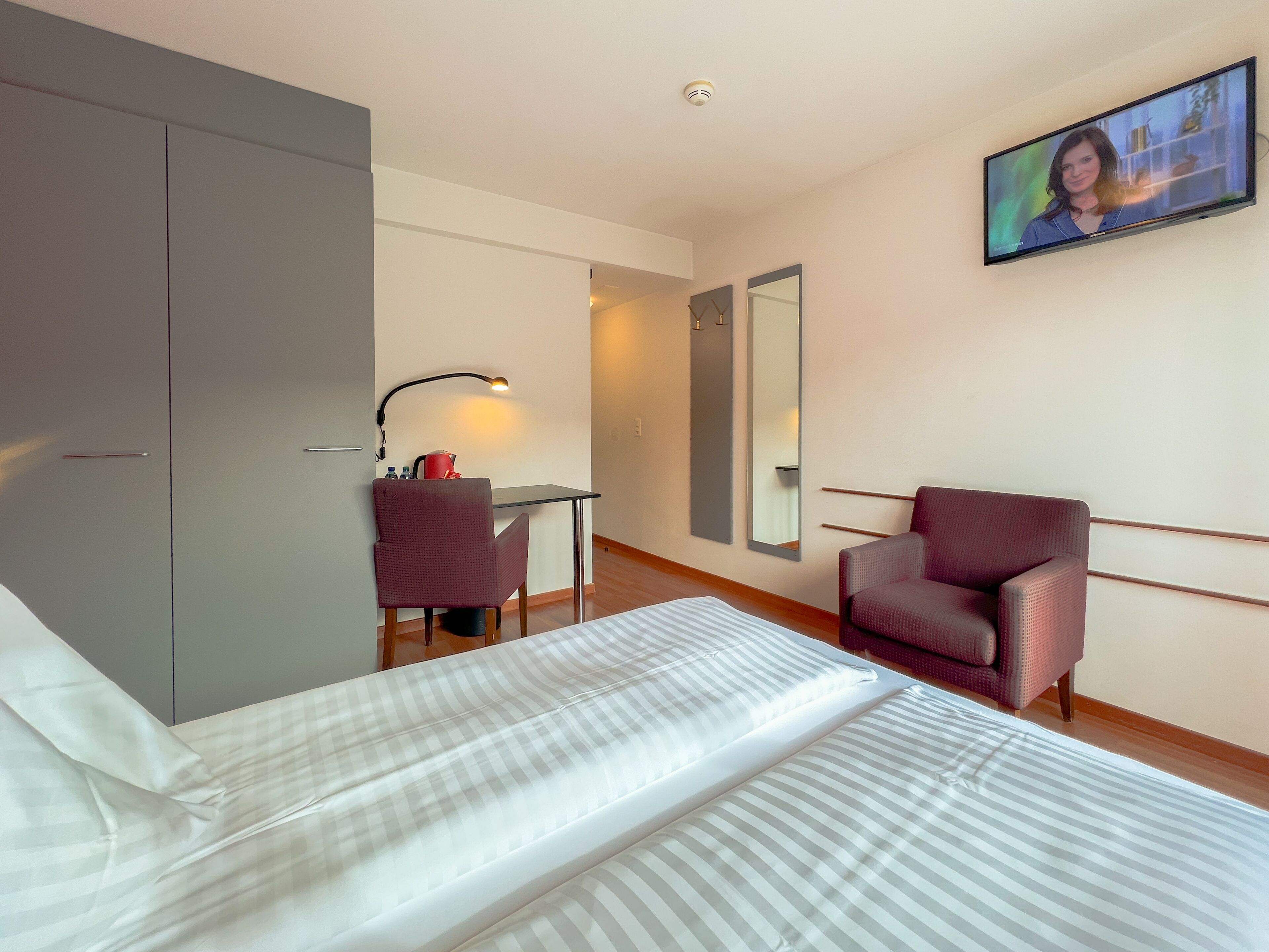 A hotel room with a bed, a desk with a chair, a TV, a wardrobe, and a mirror.