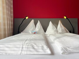 Una cama con ropa blanca y una pared roja. Hay dos lámparas negras en el cabecero.