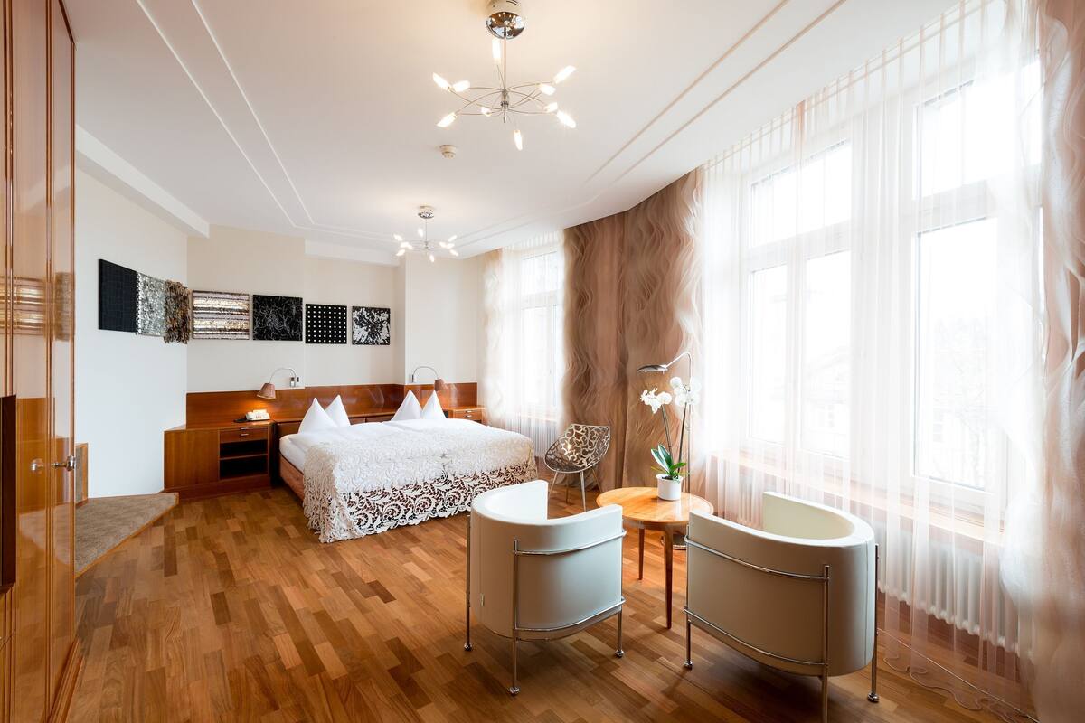 A modern hotel room with a wooden floor, a large bed with white bedding, a sitting area with a chair and table, and a large window with sheer curtains.