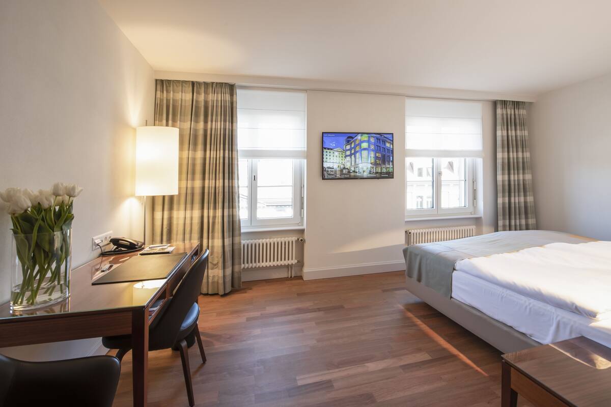A hotel room with a bed, a desk with a lamp, a chair, a vase with flowers, and a television on the wall.