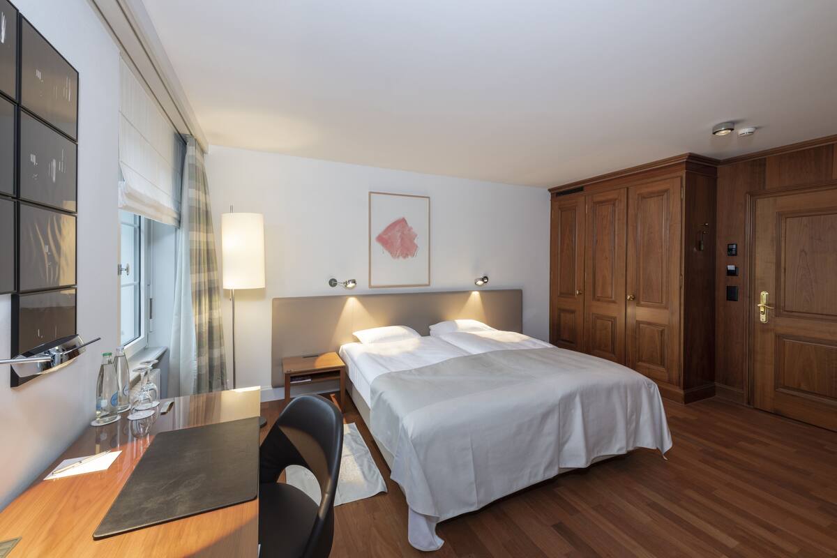 A hotel room with a bed, bedside tables, a desk, a chair, and a large window with curtains.