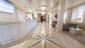 A hotel lobby with a reception desk, a chandelier, a patterned floor, and a seating area.