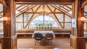 A dining area with a high wooden ceiling, a chandelier, a table set for two, and a view through a large arched window.