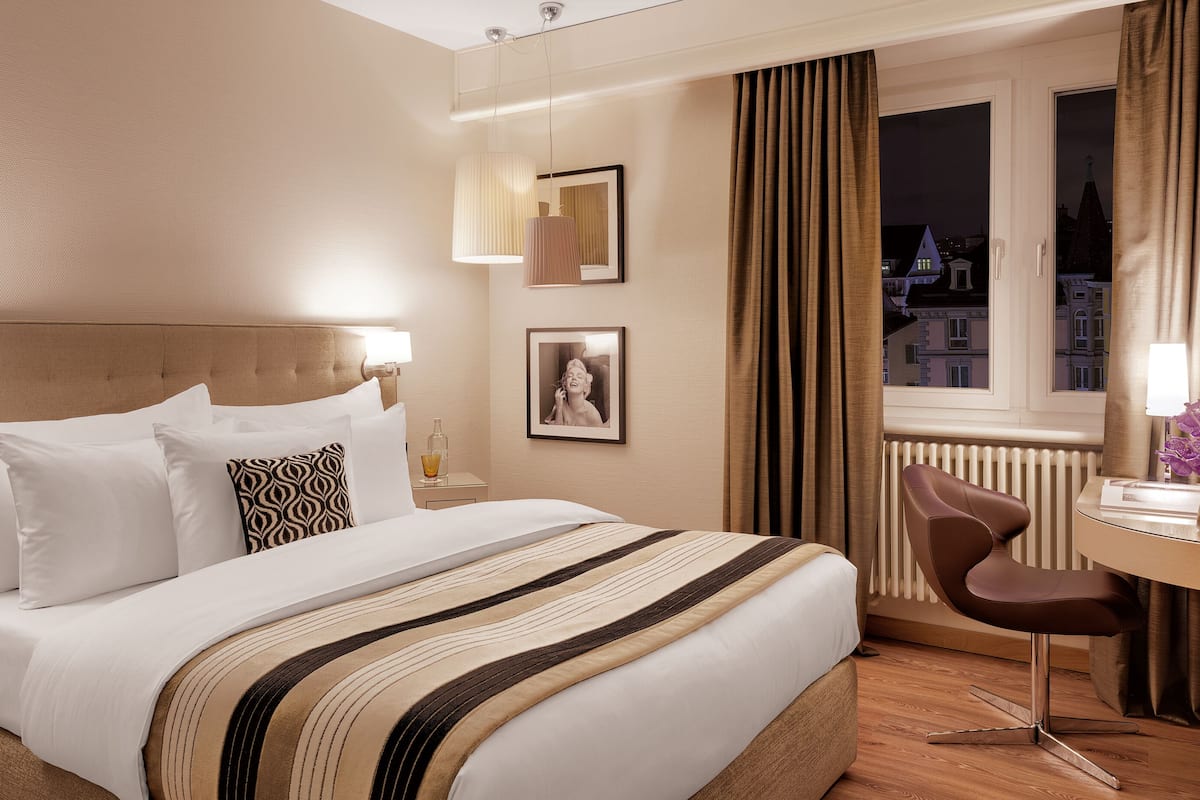 A hotel room with a bed, bedside table, lamp, chair, desk, and a TV.