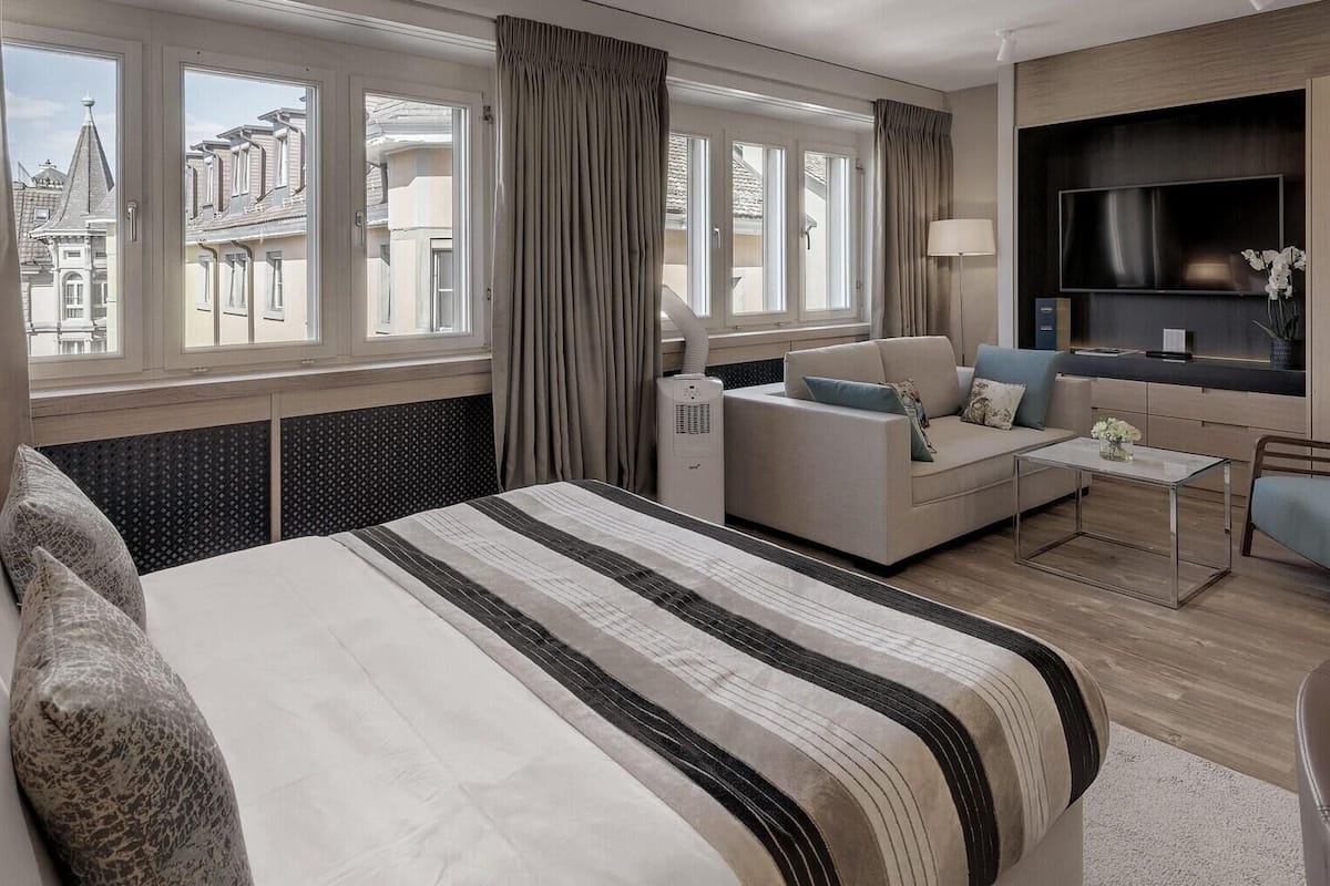 A hotel room with a large bed, a sofa, a TV, and a view of buildings outside.
