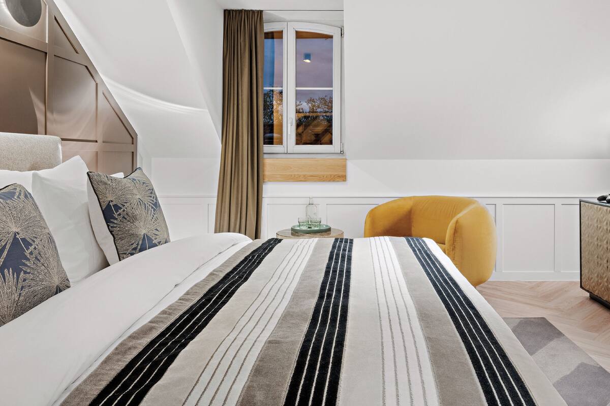 A modern bedroom with a bed, a window with curtains, a yellow chair, and a small table with a glass.