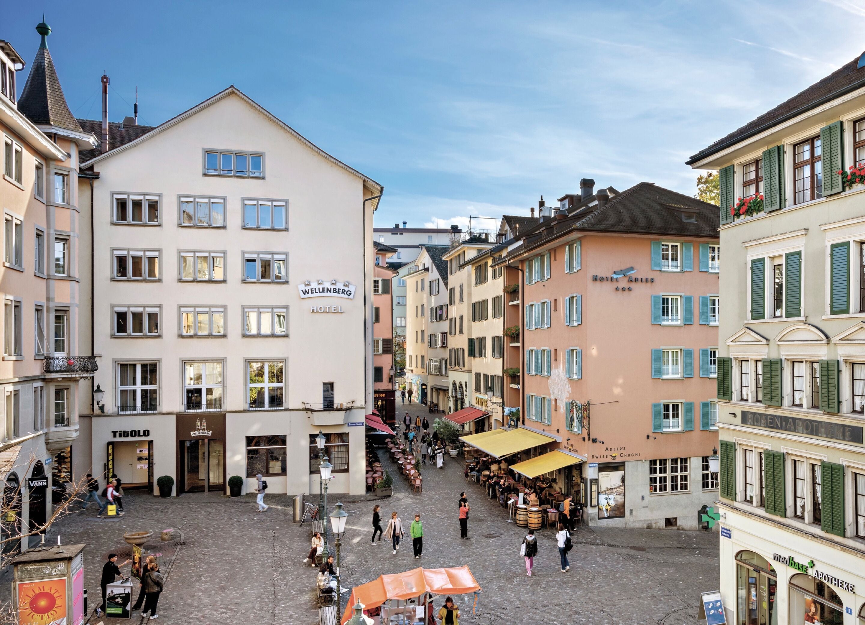 A hotel with a sign reading "Wellenberg Hotel" in a European town square with cobblestone streets and outdoor seating areas.