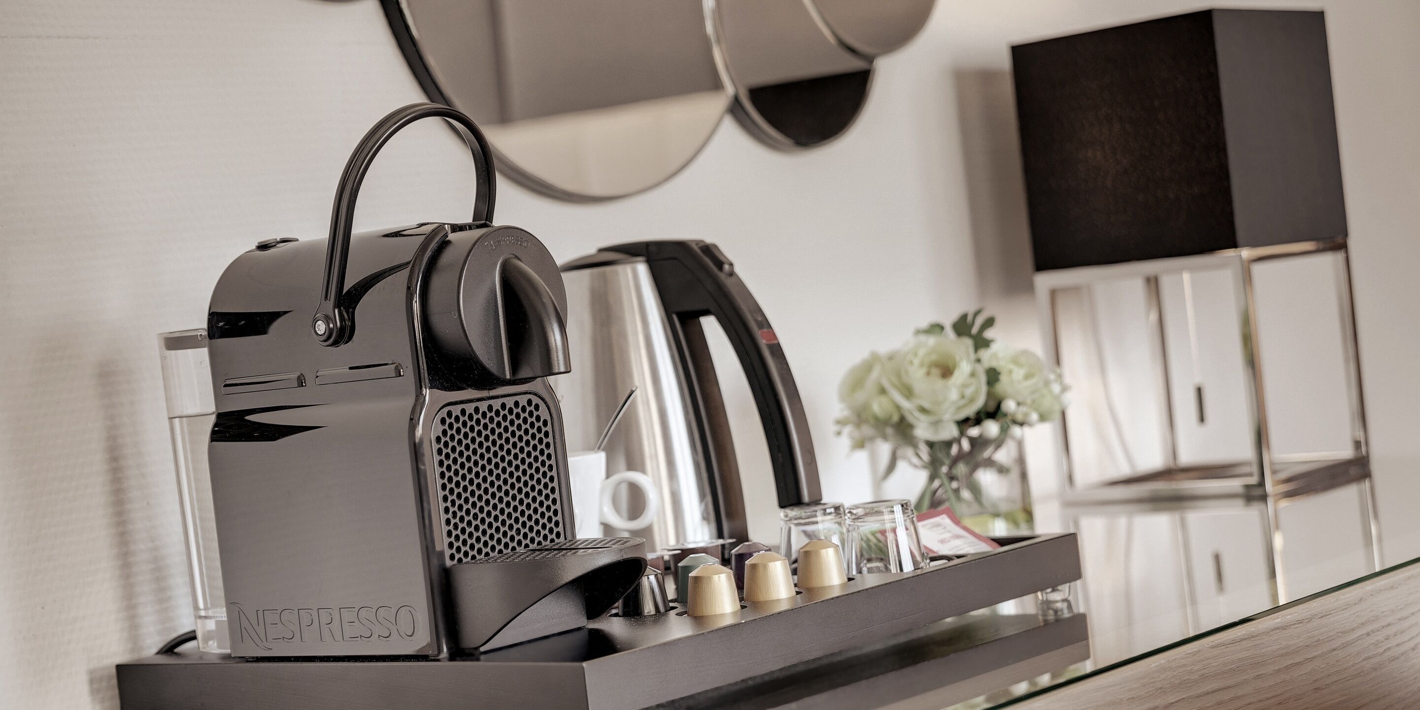 A Nespresso coffee machine on a countertop with a tray of coffee capsules and a vase of flowers in the background.