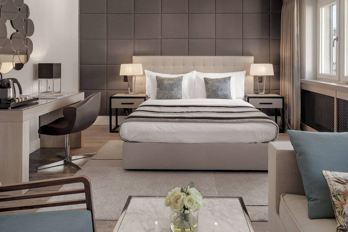 A modern hotel room with a large bed, bedside tables with lamps, a desk with a chair, a sofa, and a view of the city through the window.