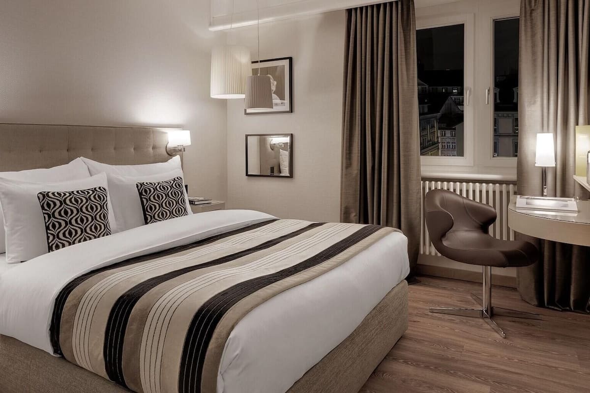 A hotel room with a bed, a desk, a chair, a mirror, and a window with curtains.