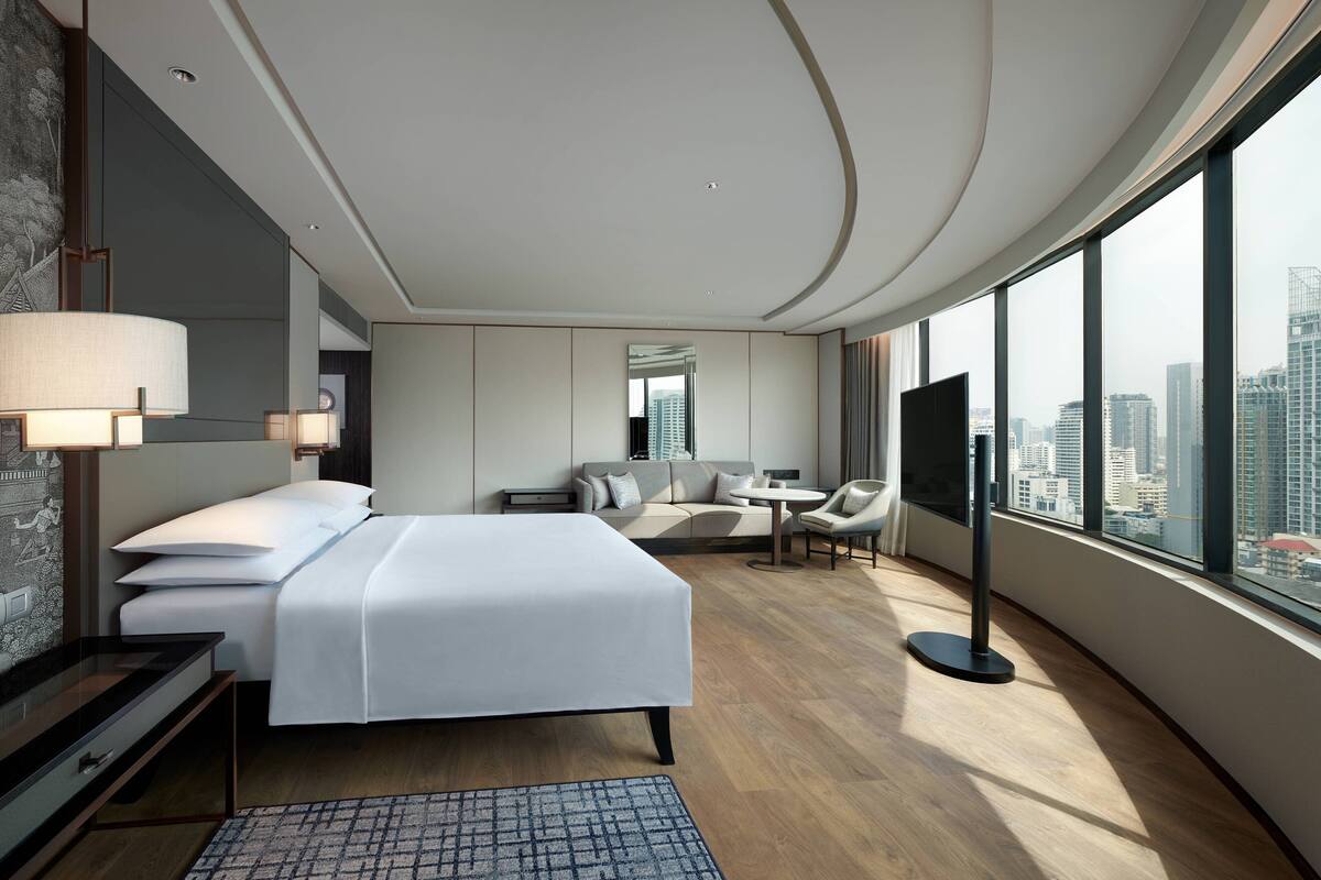 A modern hotel room with a large bed, a desk, a chair, a TV, and a view of the city.