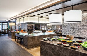 A modern hotel kitchen with marble countertops, a buffet area, and a dining area with tables and chairs.