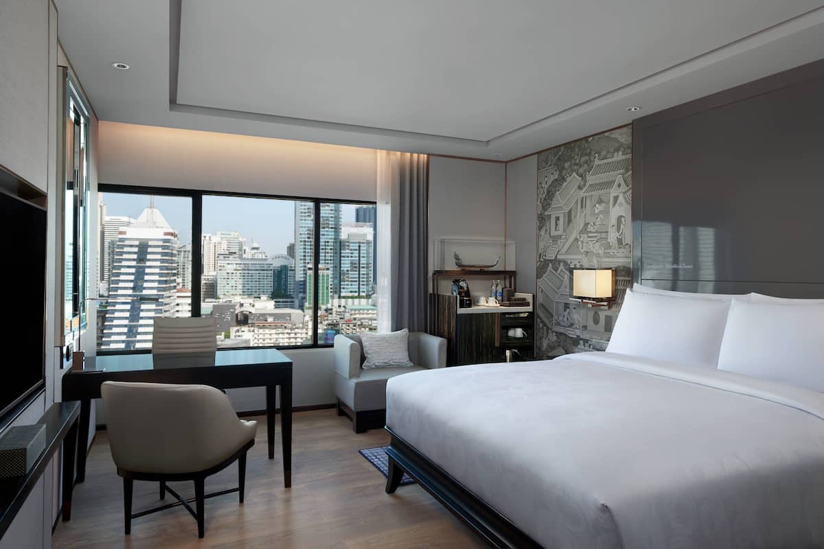 A hotel room with a large bed, a desk, a chair, and a view of the cityscape through large windows.