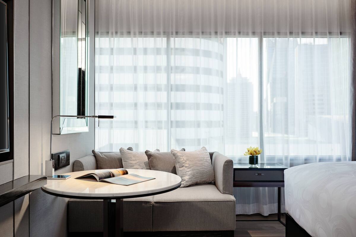 A modern hotel room with a sofa, a small round table, a bedside table, and a large window with sheer curtains.