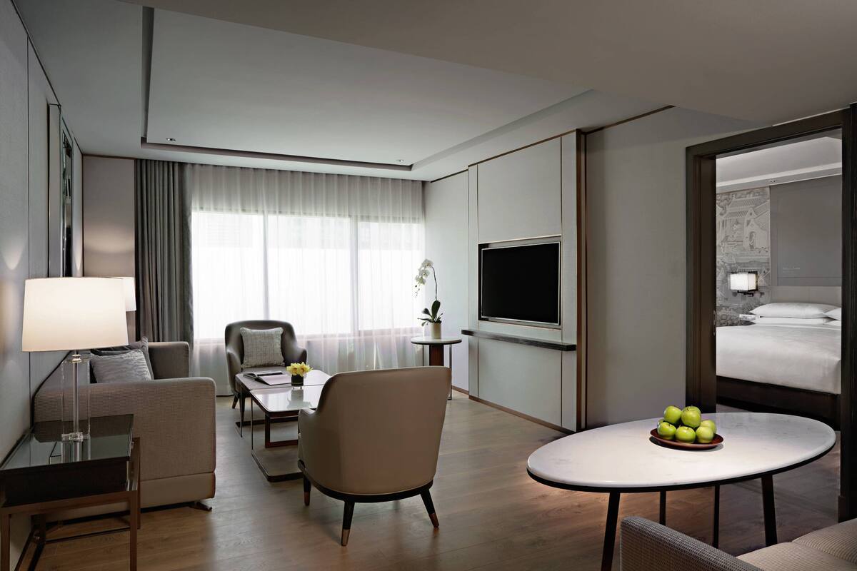 A modern hotel room with a sofa, armchair, dining table, and a TV. There is a bed with a view of a traditional building.