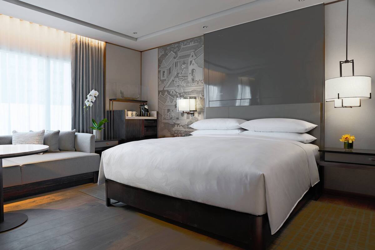 A modern hotel room with a large bed, a seating area with a table, a nightstand with a lamp, and a decorative wall panel.