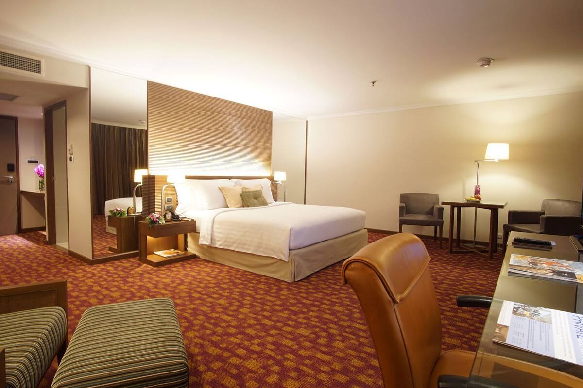 A hotel room with a large bed, a desk with a chair, a small table with a lamp, and a seating area with two chairs.