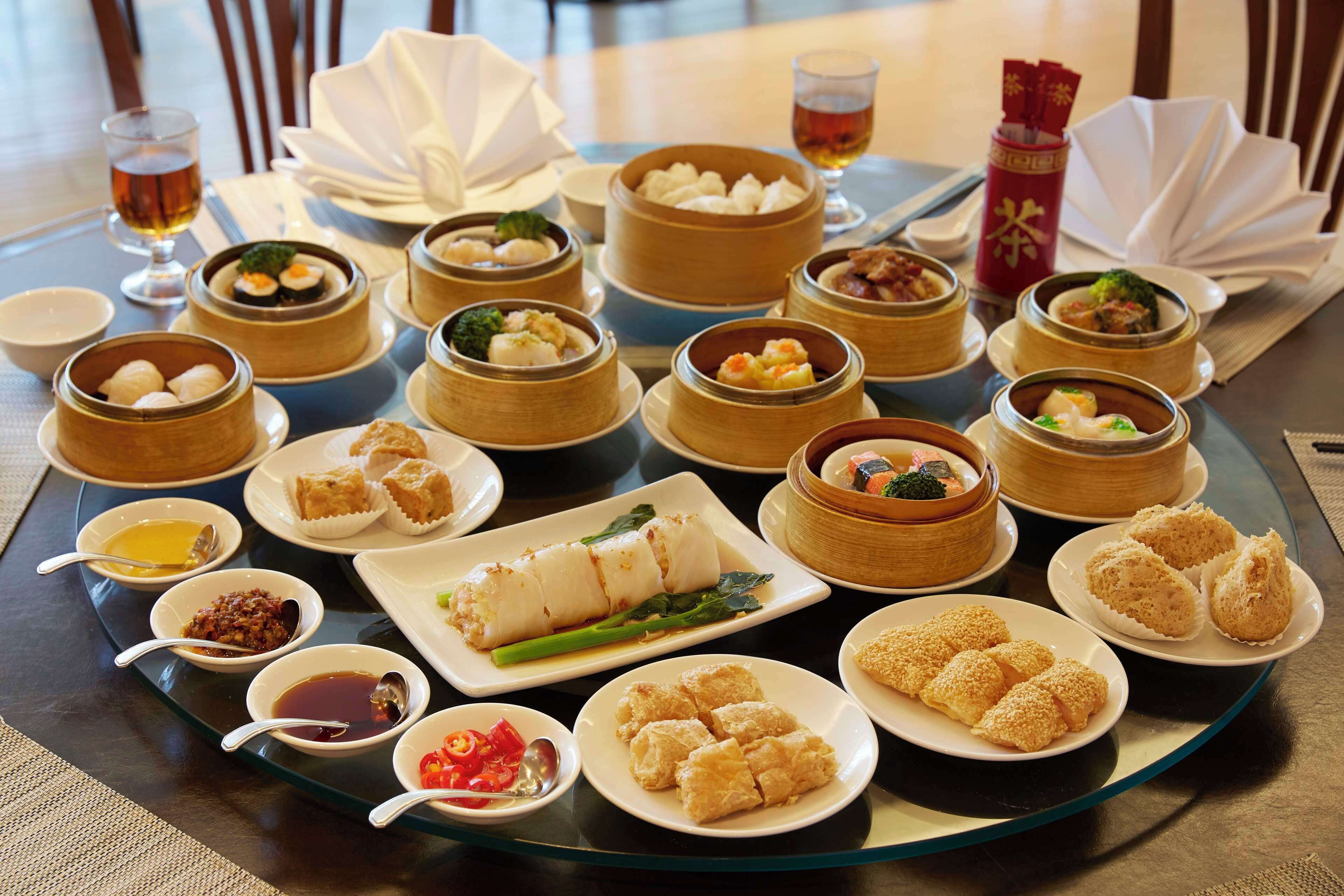 A variety of Asian dishes on a table, including dim sum and other small plates, with a glass of tea and a bottle of sauce.