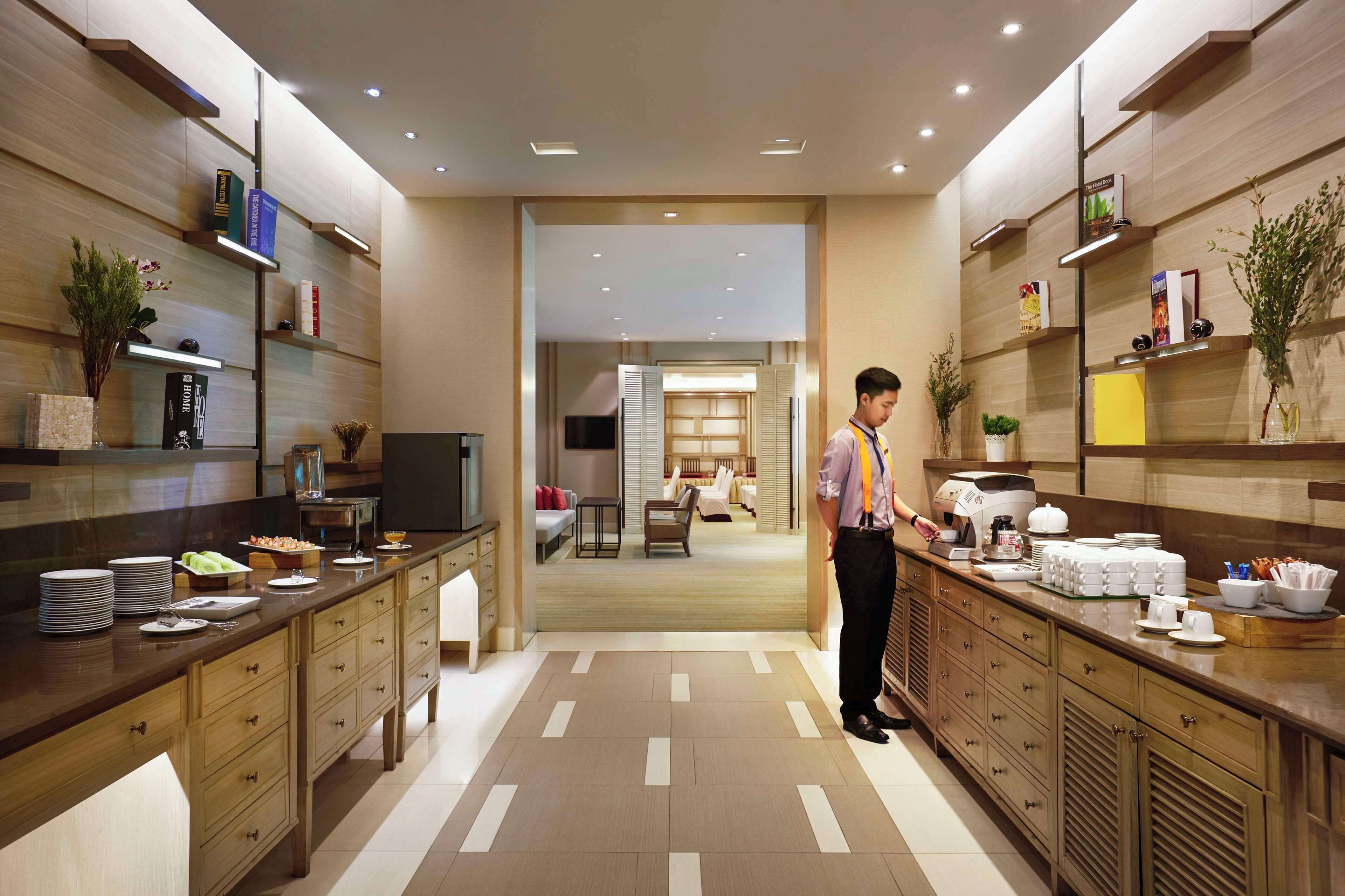 A modern hotel kitchen with wooden cabinetry, a central island, and a dining area visible through an open door.